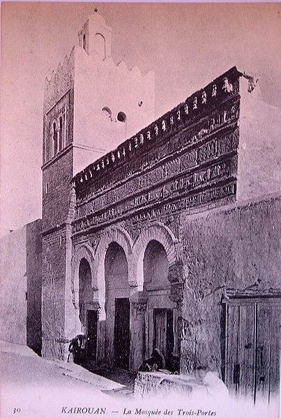 A historical black-and-white photograph of the Mosque of Three Doors in Kairouan, capturing its iconic stone façade and architectural details before modern renovations.