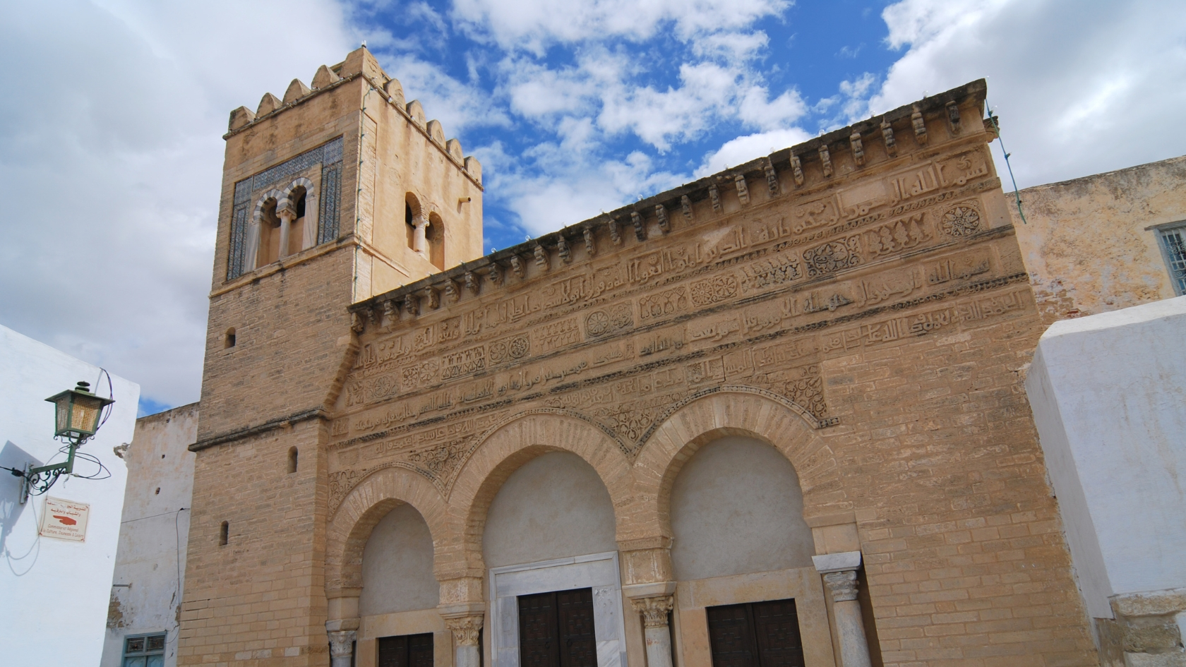 The ornate stone façade of the Mosque of Three Doors in Kairouan, showcasing three horseshoe-arched doors framed by intricate floral carvings and Kufic inscriptions